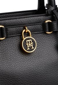 Crna kožna torbica s teksturiranim finišem, opremljena zlatnim detaljima i istaknutim okruglim logotipom.