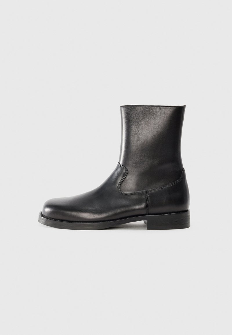 Bota de tobillo de cuero negro con una textura suave, puntera redonda, paneles laterales elásticos y una suela de goma baja texturizada.