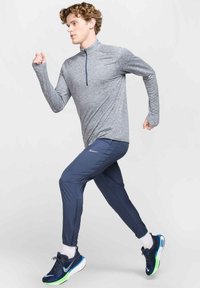 Lichtgrijze lange mouwen sporttop met ritssluiting, gecombineerd met marineblauwe slim-fit broek en donkere sportschoenen met groene accenten.