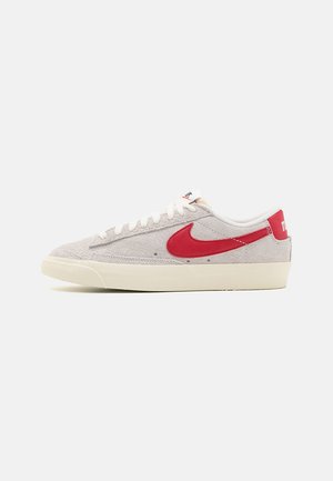 Baskets basses en daim gris clair avec le logo Nike swoosh rouge, lacets blancs et semelle en caoutchouc beige clair, présentées de profil sur fond blanc.