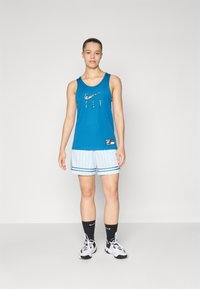 Turkos linne med texten "FLY", kombinerat med ljusblå randiga shorts. Svarta strumpor och sneakers kompletterar den atletiska outfiten.