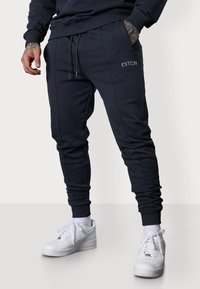 Marineblaue Jogginghose mit seitlichen Taschen, gerippten Bündchen und Kordelzugbund. Mit gesticktem Logo auf dem Oberschenkel. Kombiniert mit weißen Sneakers.