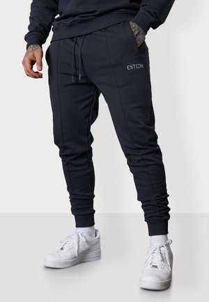 Marineblaue Jogginghose mit seitlichen Taschen, gerippten Bündchen und Kordelzugbund. Mit gesticktem Logo auf dem Oberschenkel. Kombiniert mit weißen Sneakers.
