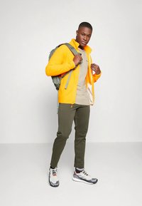 Jonge man in een felgele jas, beige shirt, olijfgroene broek, witte sneakers en met een groene rugzak op één schouder.