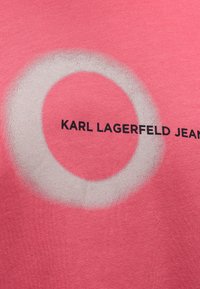 Roze tkanina s velikim bijelim kružnim uzorkom. Natpis "KARL LAGERFELD JEANS" u crnoj boji smješten unutar dizajna. Glatka tekstura.
