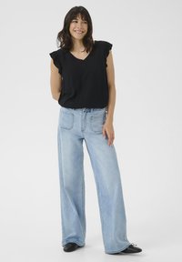 Haut noir à manches courtes avec des accents volantés, associé à un jean large bleu clair. Modèle debout, chaussures noires, arrière-plan neutre.