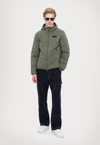 Quilted olive green jacket with a hood, zip closure, and a small logo patch; paired with dark blue pants and white sneakers.