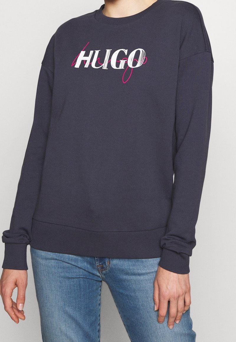 Personne portant un sweat-shirt à manches longues gris foncé avec le texte "HUGO" en blanc et rose, associé à un jean en denim bleu clair, debout contre un mur uni.