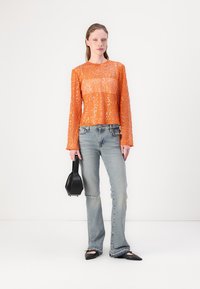 Oranje paillettentop met lange mouwen, gecombineerd met lichtblauwe flared jeans. Model houdt een kleine zwarte handtas vast. Platte zwarte schoenen maken de look compleet.