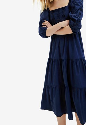 Kobieta w granatowej, długorękawowej sukience o wielowarstwowym kroju z haftowanymi kwiatowymi zdobieniami, z założonymi ramionami, stojąca na białym tle.