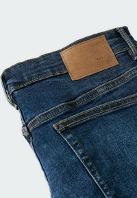 Tumma siniset farkut, joissa on teksturoitu viimeistely, kontrastiväriset oranssit ompeleet ja suorakaiteen muotoinen nahkainen merkki vyötäröllä.