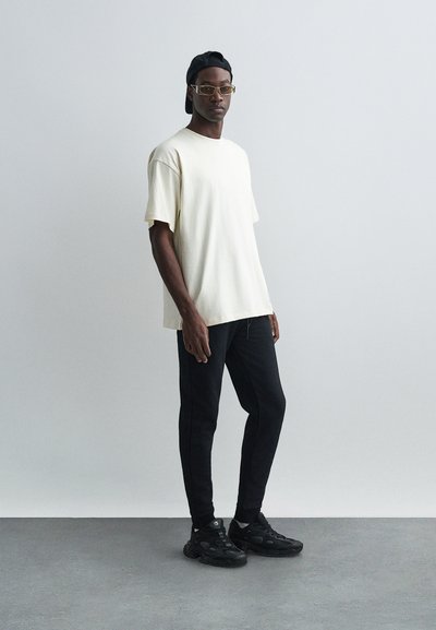 Camiseta blanca y pantalones negros con un corte ajustado. El modelo lleva una gorra negra y gafas. Las zapatillas son negras con detalles texturizados.