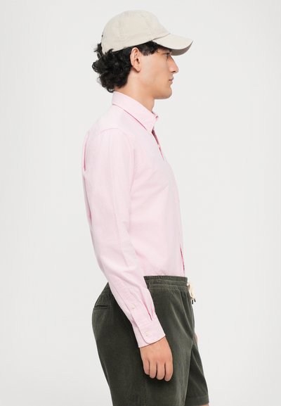 Un jeune homme portant une casquette beige, une chemise à boutons rose clair et un short en velours côtelé vert foncé se tient de profil sur fond blanc.