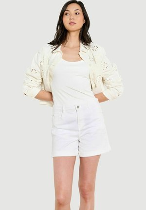 Femme portant une veste ajourée blanche sur un débardeur blanc avec un short blanc taille haute, debout, les mains dans le dos.