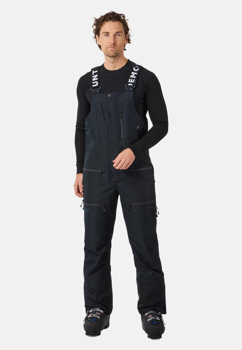 Marineblauwe skibroek met verstelbare bandjes, meerdere met rits afgesloten zakken, een gestructureerde stof en een relaxte pasvorm, gedragen over een langemouwen shirt.