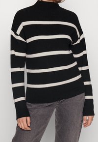 Personne portant un pull noir côtelé avec des rayures horizontales blanches et un jean gris, debout les bras détendus le long du corps.