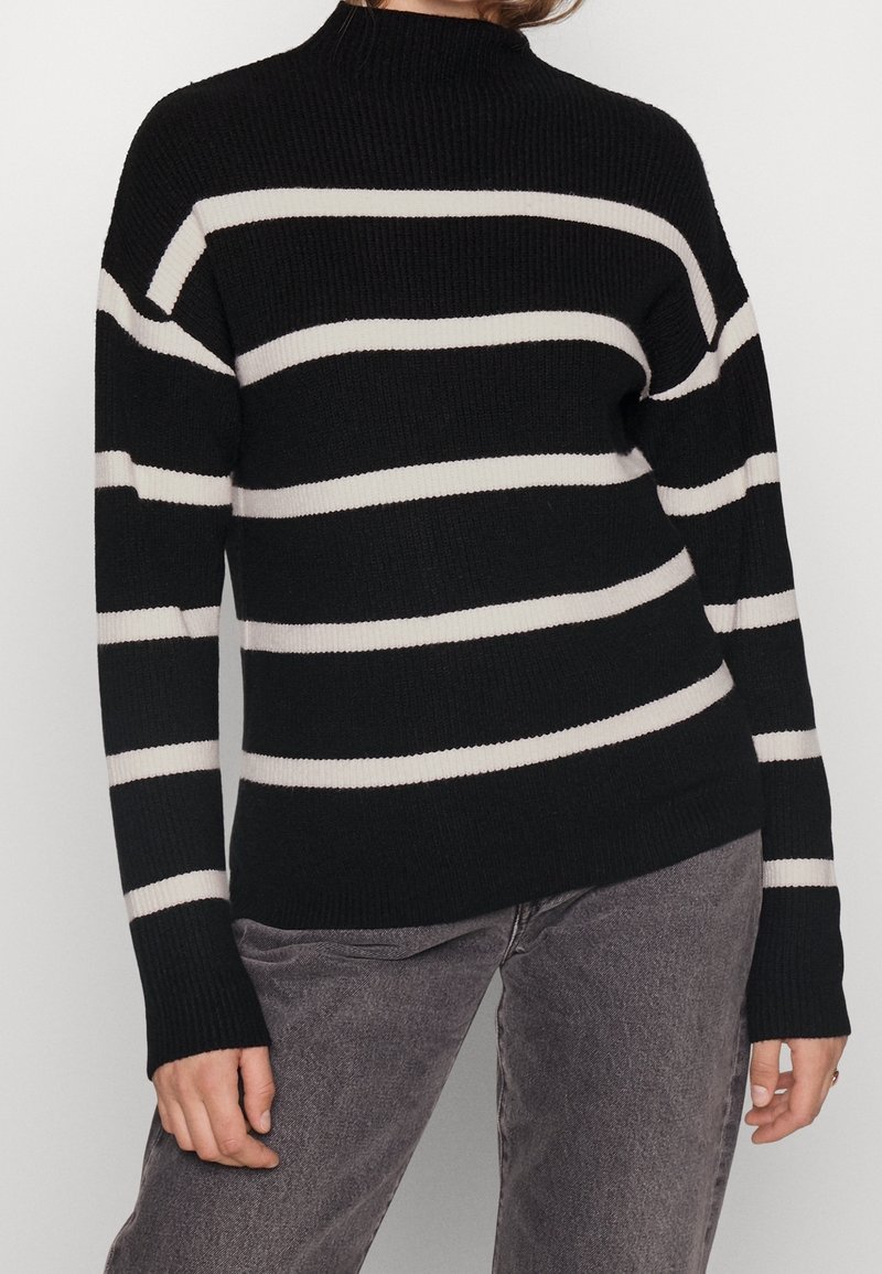 Personne portant un pull noir côtelé avec des rayures horizontales blanches et un jean gris, debout les bras détendus le long du corps.