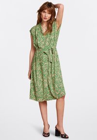 Abito floreale verde con scollatura a V e maniche corte, caratterizzato da una vita con laccetto e un tessuto morbido e fluido. Tacchi aperti neri completano il look.