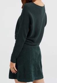 Maglione verde scuro a coste con schiena scoperta, abbinato a una morbida gonna verde scamosciato. L'outfit è caratterizzato da una vestibilità comoda e tessuti testurizzati.