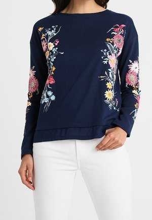 Kobieta ubrana w granatowy top z długim rękawem z wielokolorowymi kwiatowymi nadrukami z przodu i na rękawach, w połączeniu z białymi spodniami, ręką opartą na biodrze.