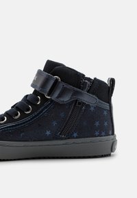 Zapatillas altas de color azul marino con patrones de estrellas, parte superior de tela, detalles de cuero, cierre con cremallera y suela de goma. Cuenta con cordones y una correa de velcro.