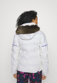 Weißes Daunenjacke mit einem von Fell gefütterten Kapuze, mit blauen Nähten und einer strukturierten Optik. Kombiniert mit floral gemusterten Hosen.