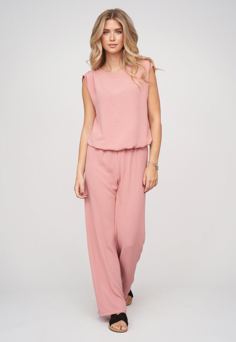 Femme aux longs cheveux blonds portant un haut rose sans manches et un pantalon large rose, marchant vers l'avant sur un fond uni clair.