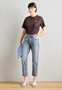 Braunes T-Shirt mit kurzen Ärmeln und blauer bestickter Detail, hoch taillierte hellblaue Jeans und hellblaue Slipper.