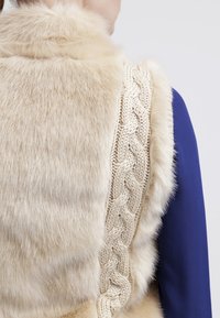 Personne portant une veste en fourrure beige avec des détails torsadés en tricot sur un haut bleu à manches longues, vue de dos et au niveau de l'épaule.