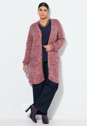 Femme portant un long cardigan en tricot violet, un haut marine avec un décolleté découpé, un pantalon large foncé et des bottes à talons violettes, se tenant contre un mur blanc.