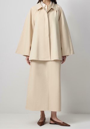 Kvinde iført beige oversized jakke og matchende lang nederdel, kombineret med brune spidse slingback-flats på neutral baggrund.