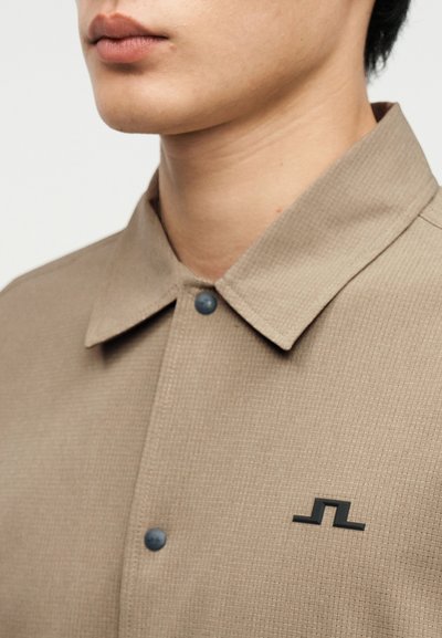 Κοντινή λήψη ενός ατόμου που φοράει μια μπεζ πουκάμισο με γιακά, μαύρα κουμπιά τύπου snap και ένα μικρό μαύρο γεωμετρικό λογότυπο στο αριστερό στήθος.