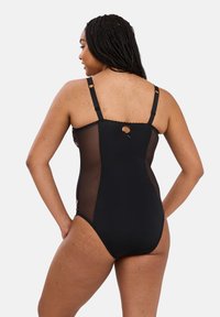 Femme aux cheveux tressés portant un body noir à panneaux transparents vu de dos, se tenant contre un fond clair.