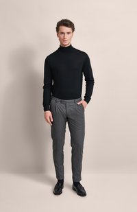 Schwarzer Rollkragenpullover kombiniert mit grauen, gefalteten Hosen und schwarzen Slippern, stehend vor einem neutralen Hintergrund.