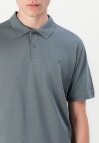 Grå polo-shirt lavet af tekstureret stof. Har en klassisk krave, to knapper og et lille logo på venstre side. Korte ærmer.