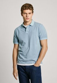 Light blue polo shirt with a contrasting collar, short sleeves, and a subtle logo. Worn over a white T-shirt, paired with dark jeans.