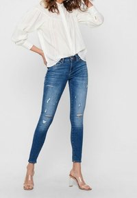 Lättviktig vit skjorta med lång ärm som bärs tillsammans med åtsittande blå denimjeans med slitage och rivna detaljer. Klara klacksandaler.