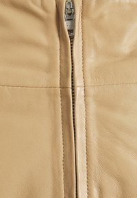Giacca di pelle beige con zip centrale, hardware YKK, texture liscia, dettaglio delle cuciture frontali e superficie leggermente venata.