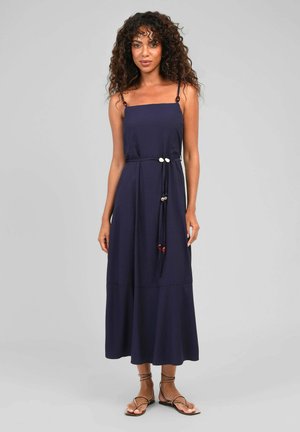Femme aux cheveux bouclés portant une robe longue sans manches bleu marine avec une ceinture perlée et des sandales marron à brides, debout devant un fond uni.