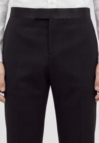 Gros plan sur un pantalon noir de costume porté avec une chemise blanche, montrant la taille, la ceinture et la fermeture par bouton.