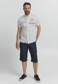 Camisa blanca de botones de manga corta con detalles impresos, combinada con pantalones cortos cargo azul oscuro y zapatillas negras. Conjunto sencillo y casual.