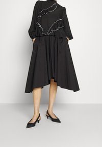 Svart klänning med rynkdetaljer, korta ärmar och fickor. Matcher med svarta slingback-klackar. Slät textur, knähög fåll.