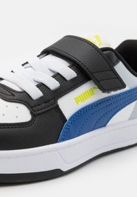 Baskets noires et blanches avec accent bleu, en matériau synthétique en cuir, semelle plate, fermeture à lacets et à sangle, comportant un détail de logo jaune.