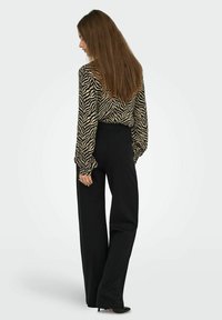 Donna in posa laterale che indossa una blusa a maniche lunghe con stampa zebra, pantaloni neri a vita alta e ampia gamba, e tacchi neri.