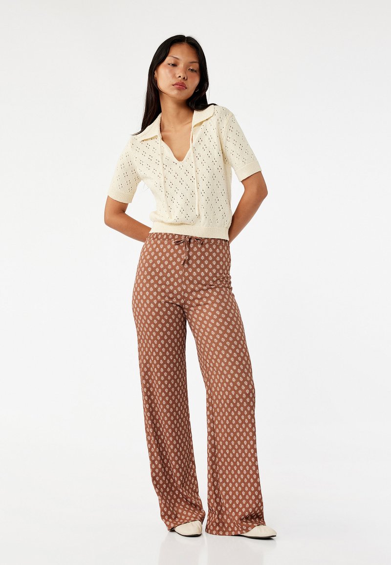 Strickpulli in Creme mit kurzen Ärmeln und Lochmuster sowie V-Ausschnitt. Lockere braune gemusterte Hose mit weitem Bein und Kordelzugbund.