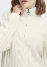 Pull en maille blanche avec un col haut et une fermeture éclair quart, présentant un motif en torsades et une texture côtelée sur les manches.