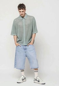 Jovem a usar uma camisa de botão de malha de mangas curtas verde claro, calções de ganga claros e longos, meias brancas com riscas e ténis brancos.