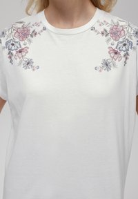 T-shirt blanc à manches courtes avec broderie florale rose, bleue et verte sur les épaules, col rond et texture douce.