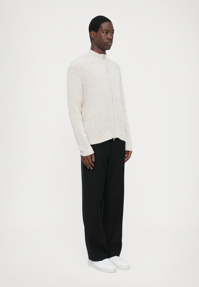Ljusgrå zip-up-tröja med hög krage, ribbad textur och långa ärmar, i kombination med svarta vida byxor och vita sneakers.