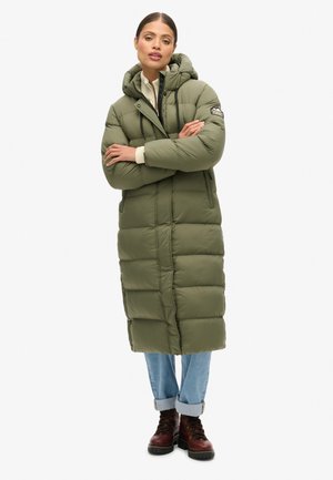 Manteau matelassé vert olive avec capuche, design matelassé, fermeture éclair à l'avant, accents de cordon et poignets côtelés, porté par-dessus un jean bleu clair et des bottes brunes.